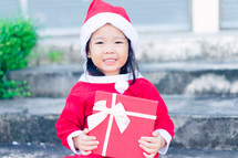 a little girl in a Santa suit holding a red gift box 
