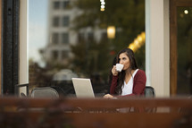 a young woman sitting outdoors at a table with a laptop sipping coffee.