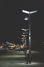 A woman illuminated by a street light while leaning against a light pole at night.