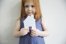 A little girl in a blue dress holds a paper cutout of a house.