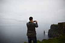 a man standing at the edge of a cliff taking a picture of the ocean 