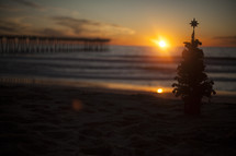 Christams tree on beach with waves and pier in background at sunset.