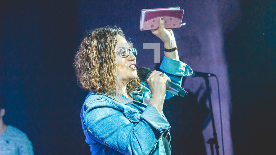 a woman holding a microphone and a raised Bible during a worship service 
