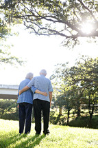 An elderly couple stand outdoors with their arms around each other.