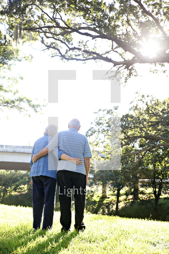 An elderly couple stand outdoors with their arms around each other.