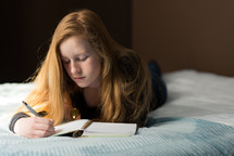 teen girl writing in a journal lying on a bed 