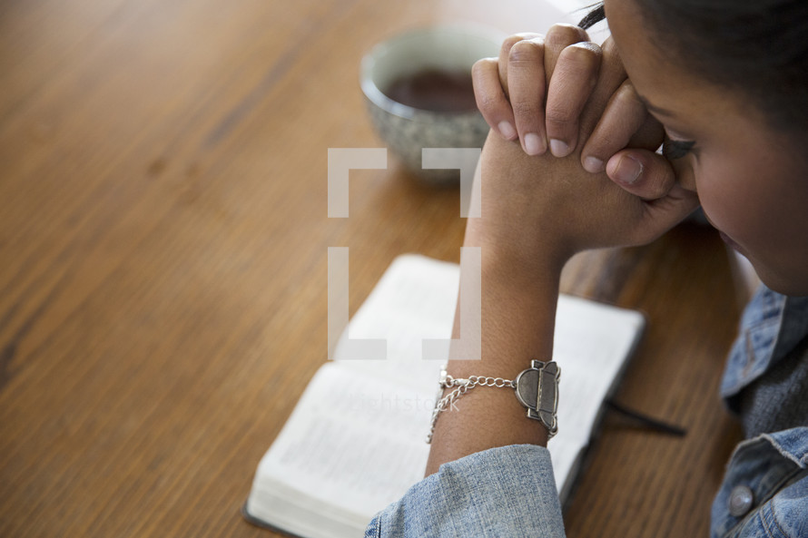 an African American woman sitting at a table reading a Bible and praying 