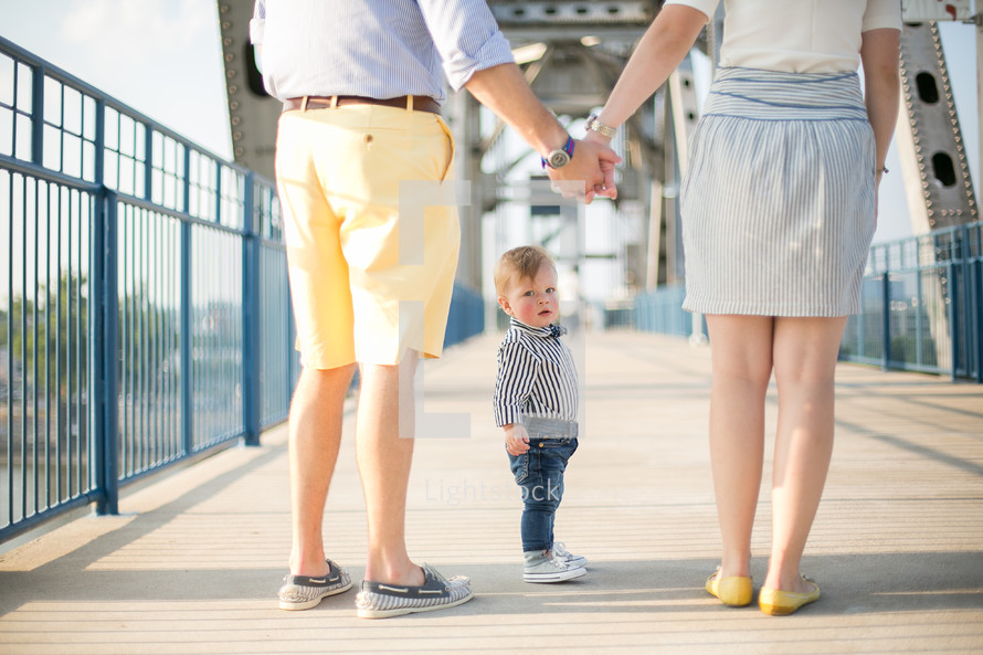 Toddler boy in a bow tie standing on a bridge with his parents who are holding hands.