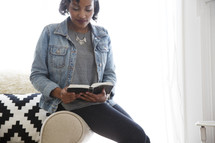 An African American woman sitting on the arm of a chair reading a Bible 