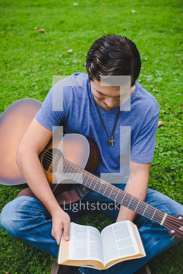young man sitting in grass playing a guitar and reading a Bible 
