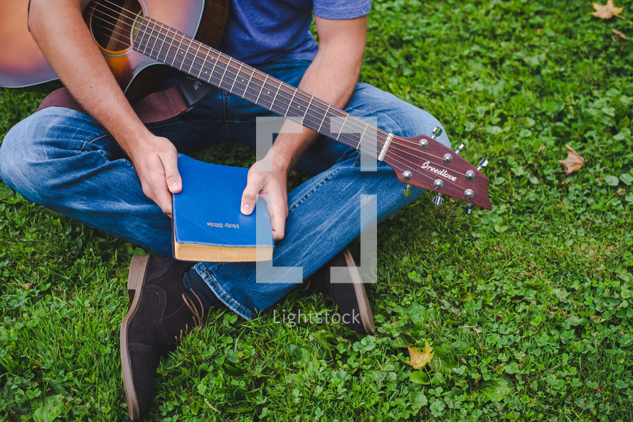young man sitting in grass playing a guitar and holding a Bible 