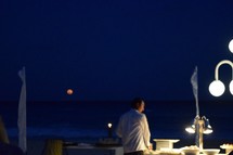A server attending to a buffet table at dusk with a view of the full moon over the ocean 