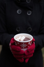 A Christmas cup of hot chocolate being held by a woman wearing red mittens.