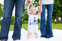 Two adults in denim jeans holding  the hands of a toddler boy standing on a white blanket in the grass in a park.