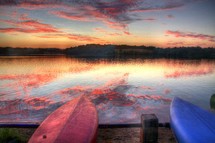 pink and purple clouds reflecting on lake water in front of two flipped over  boats resting on a shore