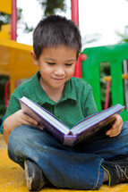 An Asian boy reading a book on a playground 