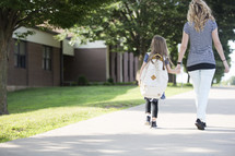 A mother and daughter holding hands and walking to school together.