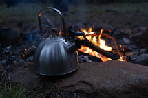 A tea kettle next to a camp fire at dusk