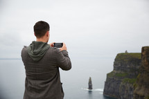 a man standing on the edge of a cliff taking a picture of the ocean 