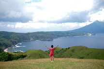 A woman in a pink dress stands on a green hill with arms outstretched toward the ocean.