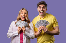 Amazed man and woman with USD currency. Young american student couple holding money - dollars banknotes on violet wall. Symbol of success, gain, salary, benefit, investments.