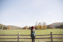 woman standing on a fence looking out at a field and mountain 