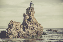 A man sitting on a rock formation in the ocean. 