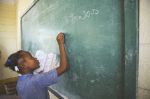 Girl holding a book while writing on a chalkboard in a classroom.