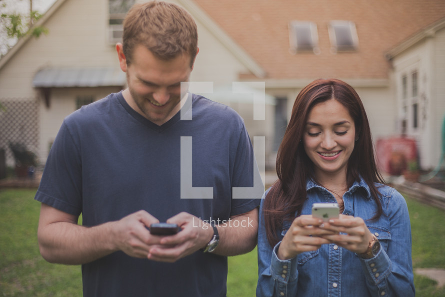 Smiling couple standing outside in front of a house texting on cell phones.