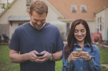 Smiling couple standing outside in front of a house texting on cell phones.