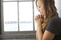 a young woman with head bowed in prayer in front of a window 
