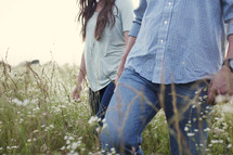 a couple walking holding hands through a field of tall grass 