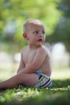 Toddler in a diaper sitting in the grass outside.