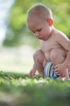 Toddler boy in a diaper squatting in the grass outside.