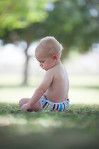 Toddler in a diaper sitting in the grass outside.