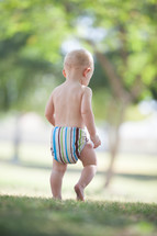 Toddler in a diaper walking in the grass outside.