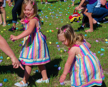 A set of twin girls with colorful dresses go Easter Egg hunting  along with other children in a green field of Easter eggs on Easter at a local church event. 
