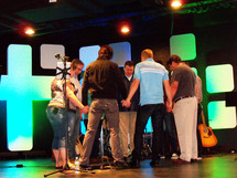 The pastors and staff at a local church gather together to have a word of prayer before the morning worship church service begins. Here, a group of seven pastors, worship leaders and musicians gather together in a circle holding hands and asking God to bless the time of worship and morning music and sermon. They are standing on a sound stage surrounded by guitars, drums, keyboards, microphone stands and lighted stage for the morning worship celebration. 
