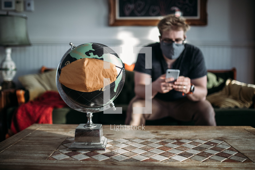 A man in a face mask looking at his cellphone near a globe with a mask 