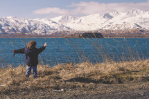 boy child standing on a lake shore looking at snow covered mountains 