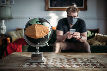 A man in a face mask looking at his cellphone near a globe with a mask 
