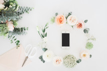 wreath of flowers around a cellphone, scissors, envelope, and bouquet of flowers 