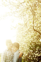 Couple embracing by flowering trees with sunbeams in background.