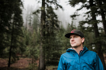 a man in a jacket and ball cap standing in a forest 
