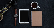 A wristwatch, cell phone, spiral notebook, cup of coffee, and a camera on a black background.