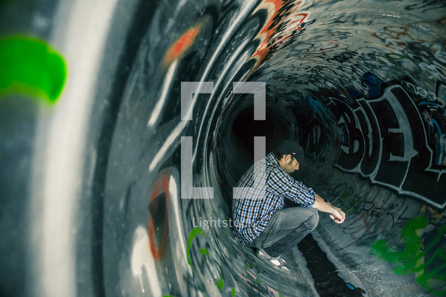 Man sitting in a sewer drain pipe painted with graffiti.