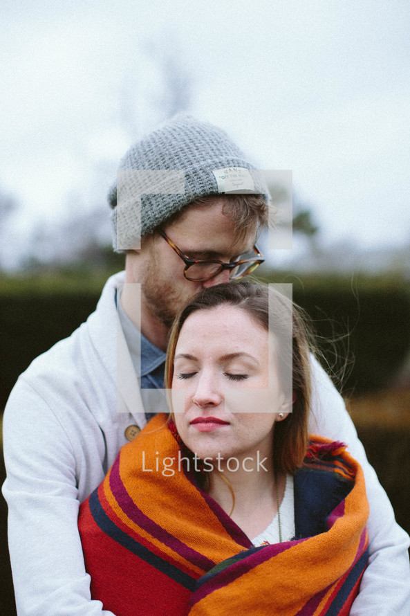 man kissing a woman wrapped in a blanket on the head 