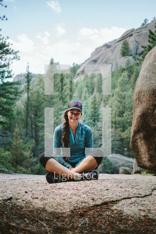 A smiling woman sitting cross legged on a boulder surrounded by a forest.