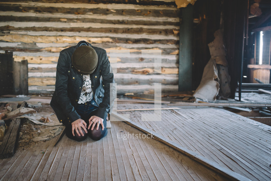A woman sits on the floor with head bent in an abandoned log house.