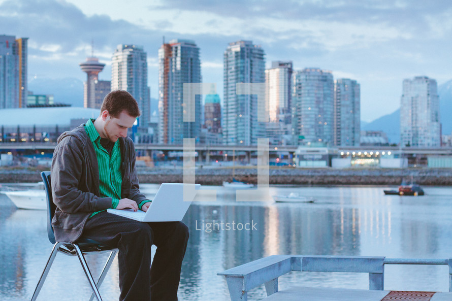 Man sitting on a chair with his laptop near an ocean bay by a city skyline.